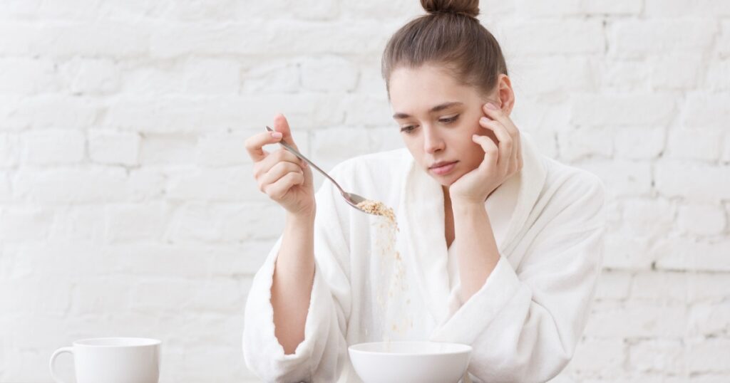 Young woman with no appetite, tired, bad mood and sleepy, sitting in the white loft kitchen, having unsavory unpalatable unappetizing breakfast