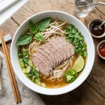 Overhead view of a steaming bowl of beef pho with rice noodles, herbs, and lime wedges